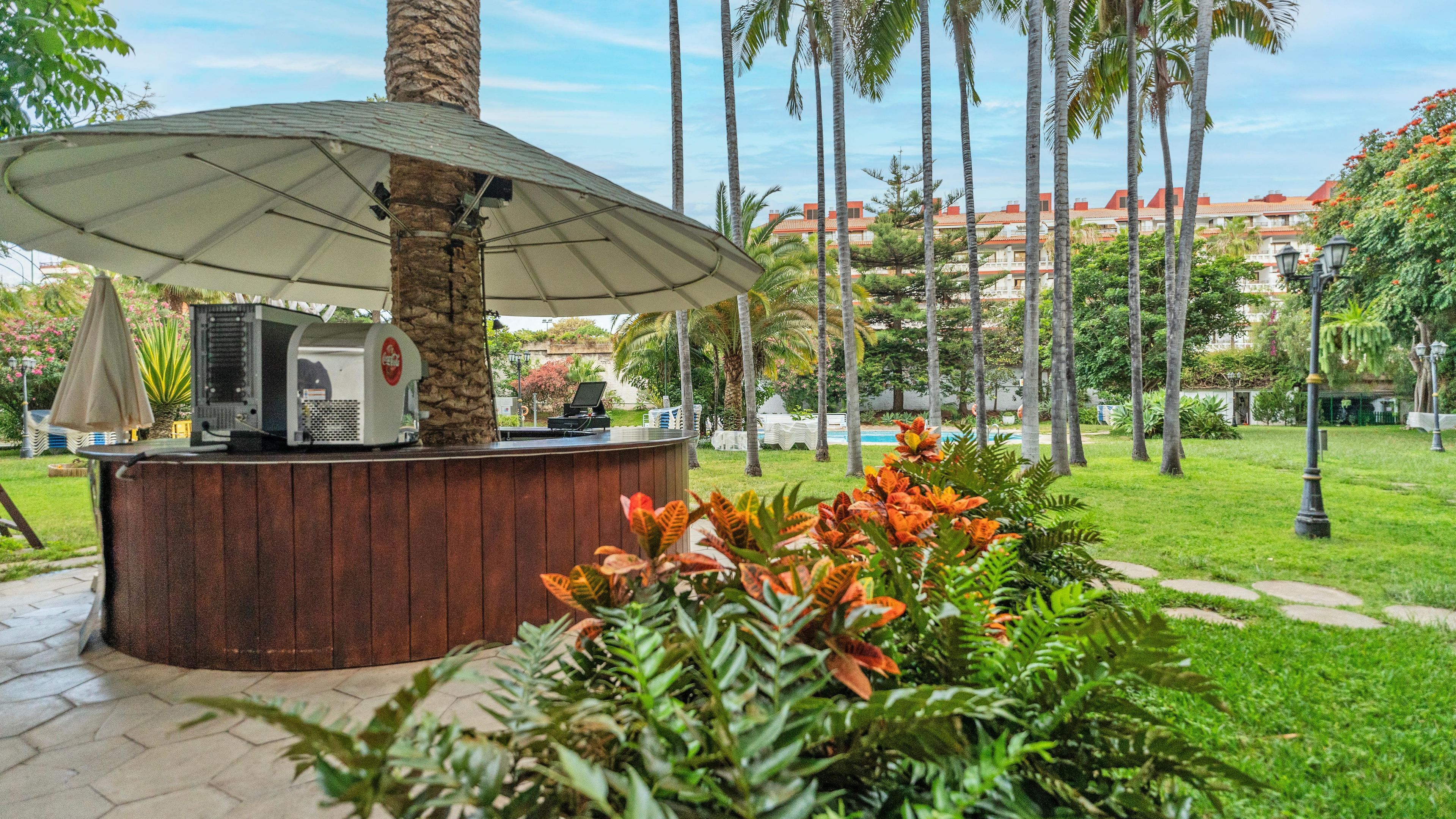 A wooden outdoor bar area with a large umbrella, surrounded by lush greenery and palm trees.