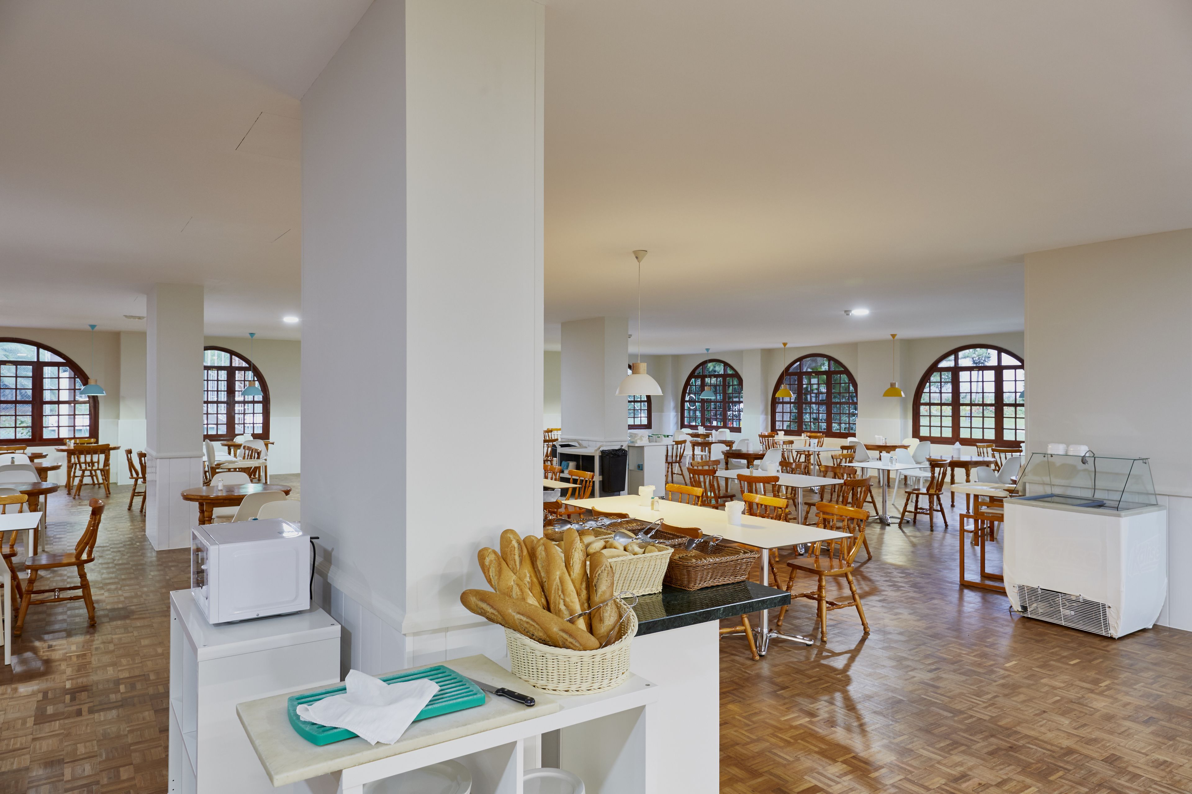 Een ruime eetruimte met meerdere tafels en stoelen, grote ramen en een buffet.