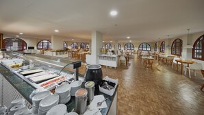 Un grand espace dédié à la salle à manger, avec de nombreuses tables et chaises, un buffet proposant une variété de plats, et de grandes fenêtres surmontées d’arcs.