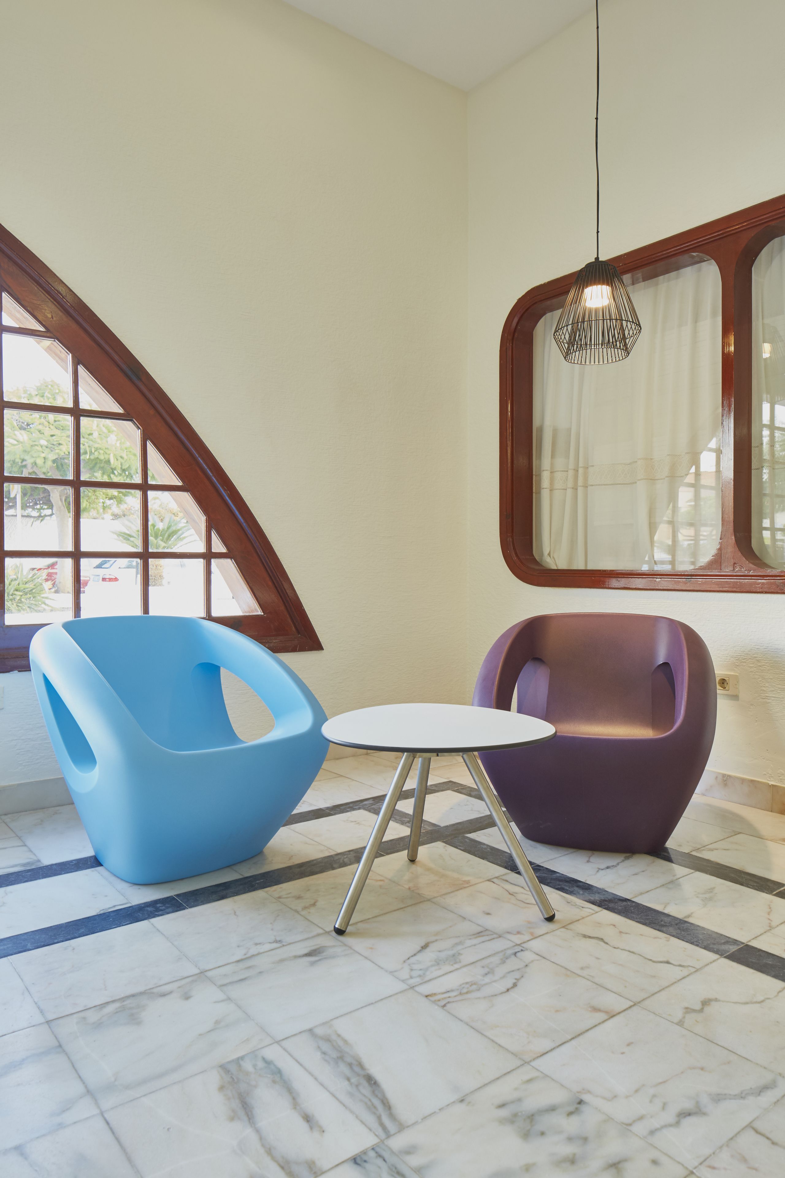Une pièce avec un sol en marbre, une chaise bleue, une chaise violette, une petite table blanche et un luminaire suspendu.