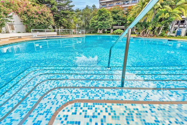 Una piscina con agua azul cristalina, rodeada de vegetación y un edificio al fondo.