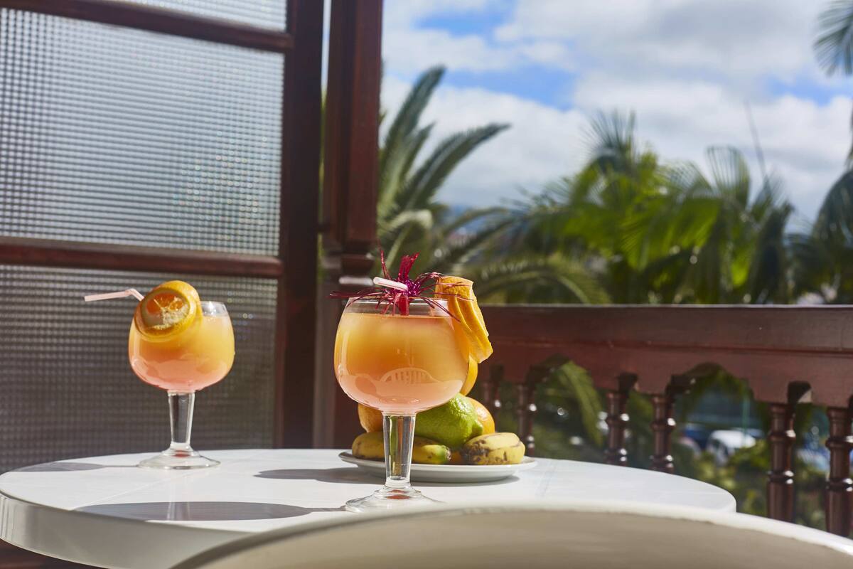 Twee cocktails op een tafel met uitzicht op palmbomen en een balkonhek.