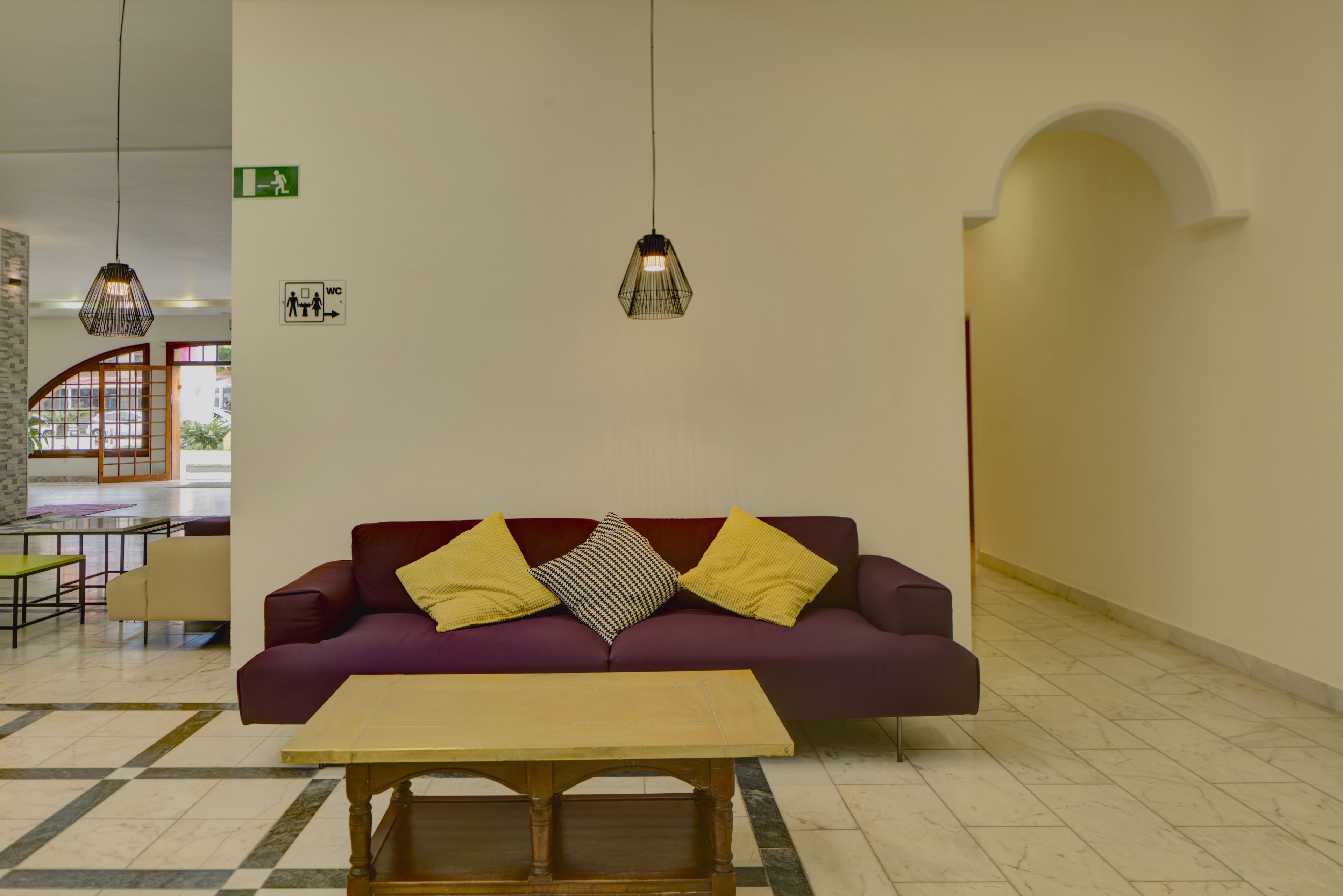 Uma área de recepção com um sofá roxo, duas almofadas amarelas, uma mesa de madeira e luminárias pendentes.