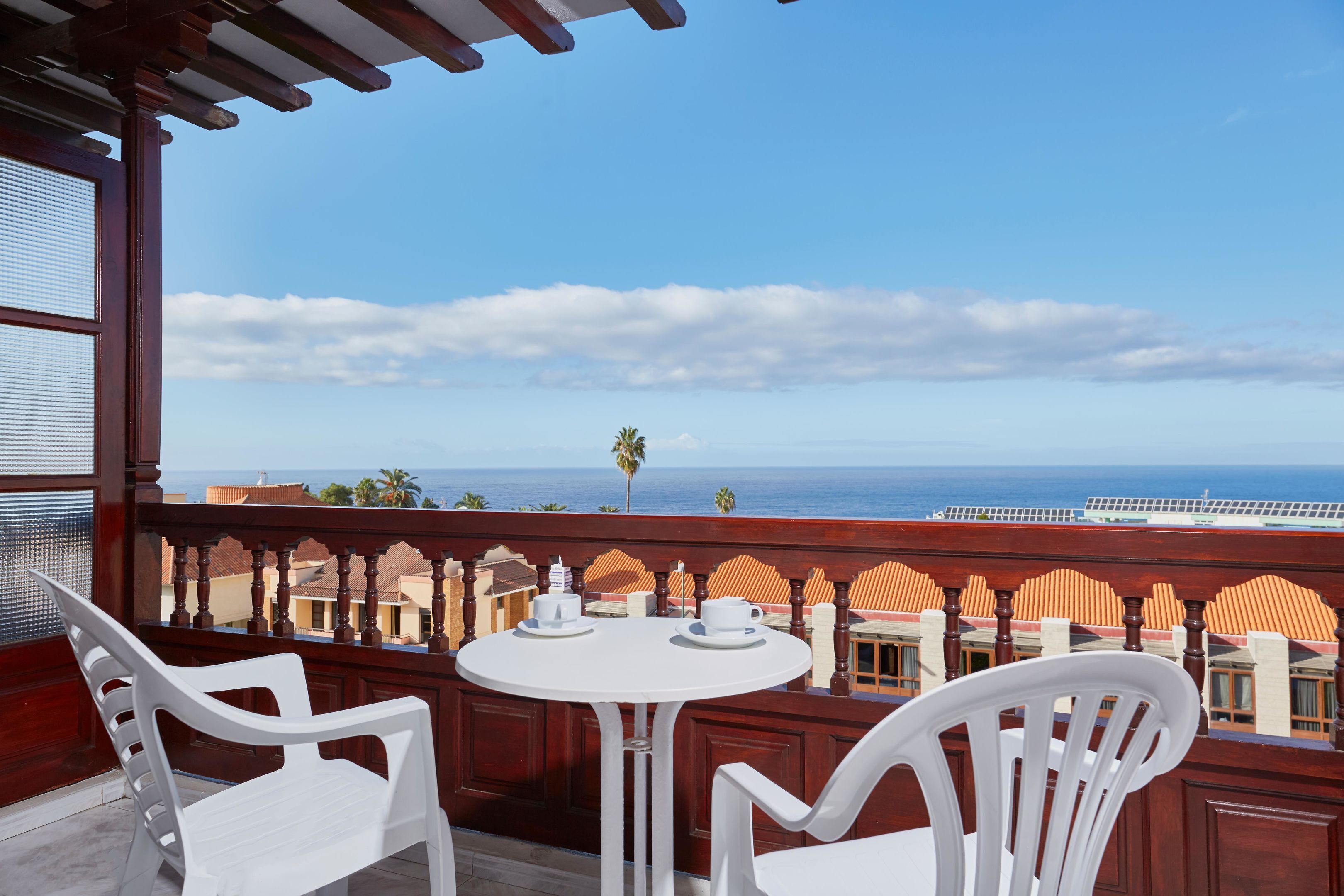 Un balcón con mesa y sillas blancas, vista al océano y una palmera.
