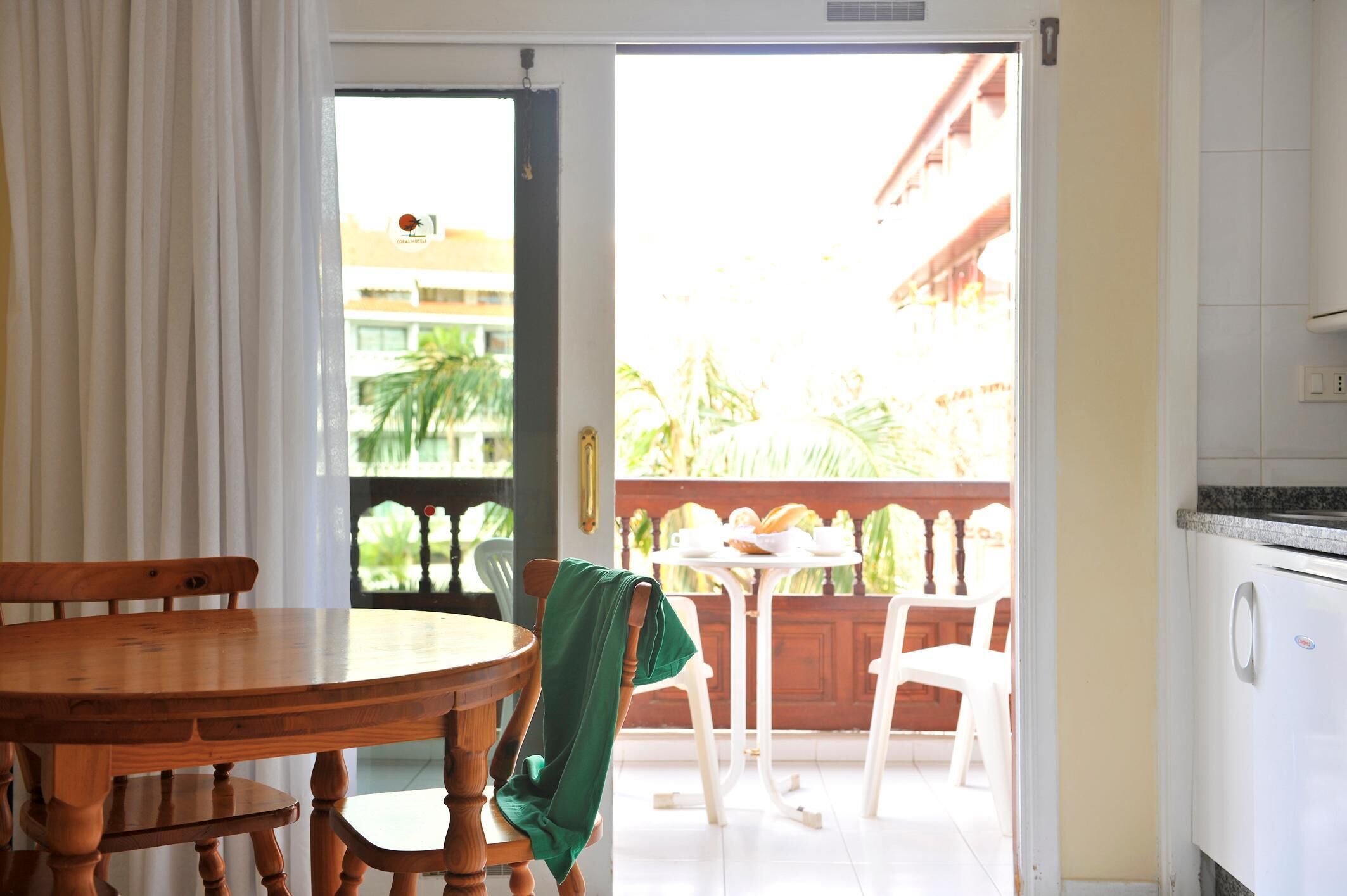 A room with a wooden dining table, a green chair, a white table, and a balcony with a view of greenery.