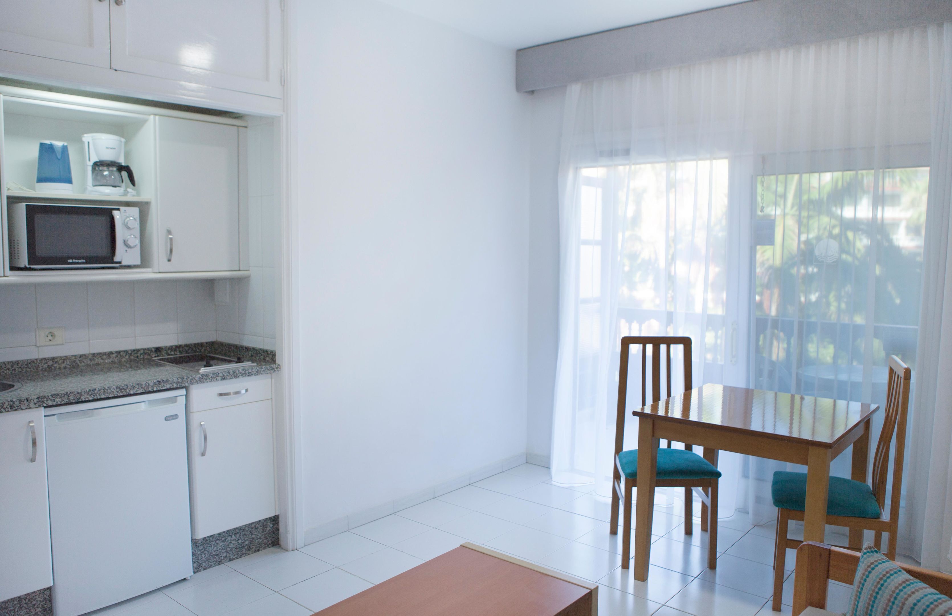 A kitchen with a microwave, coffee maker, and dishwasher. There is a dining table with chairs and a window with sheer curtains.