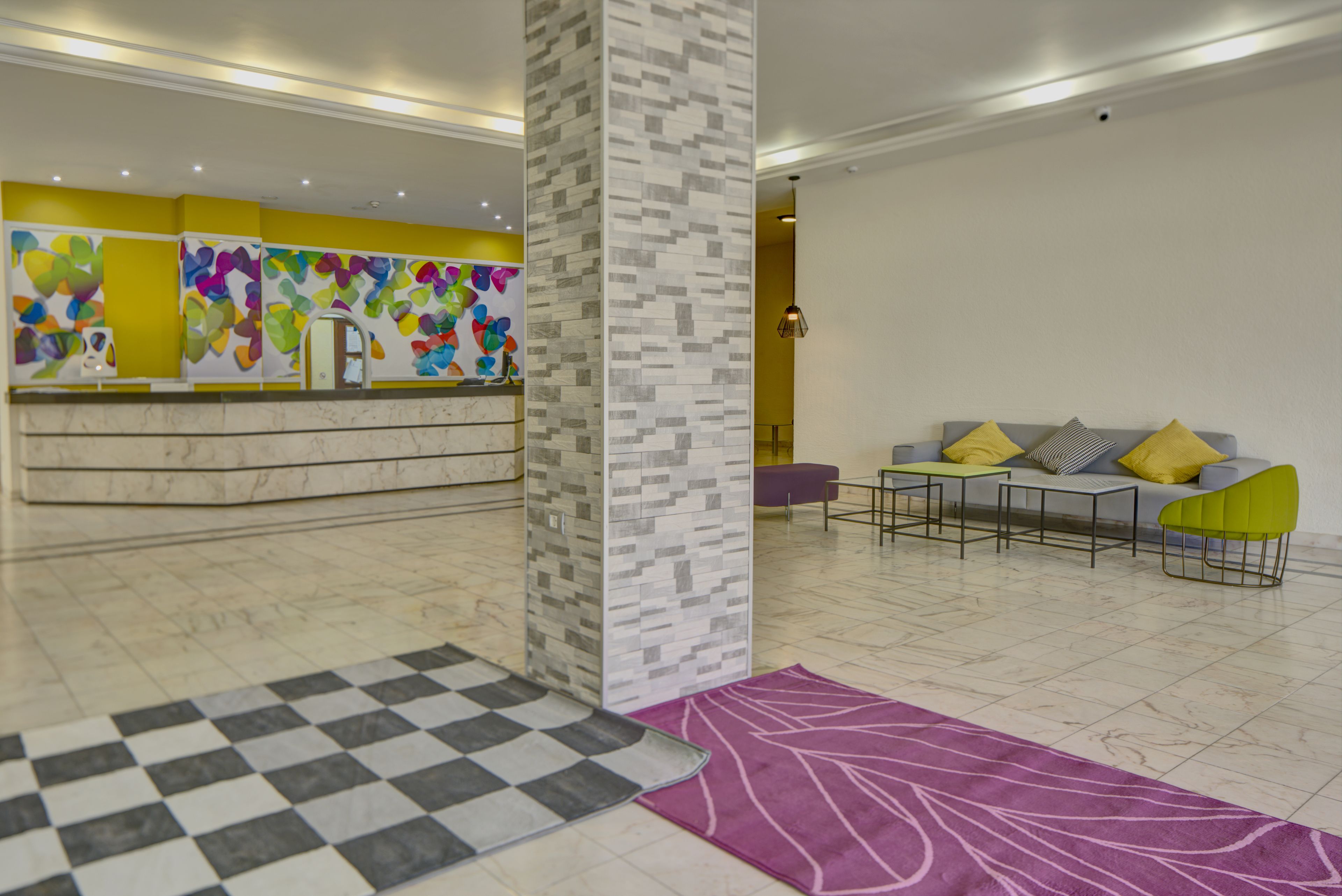 Un hall d’entrée moderne avec un comptoir d’accueil, des œuvres d’art colorées accrochées aux murs, un tapis à carreaux et un canapé agrémenté de coussins jaunes.