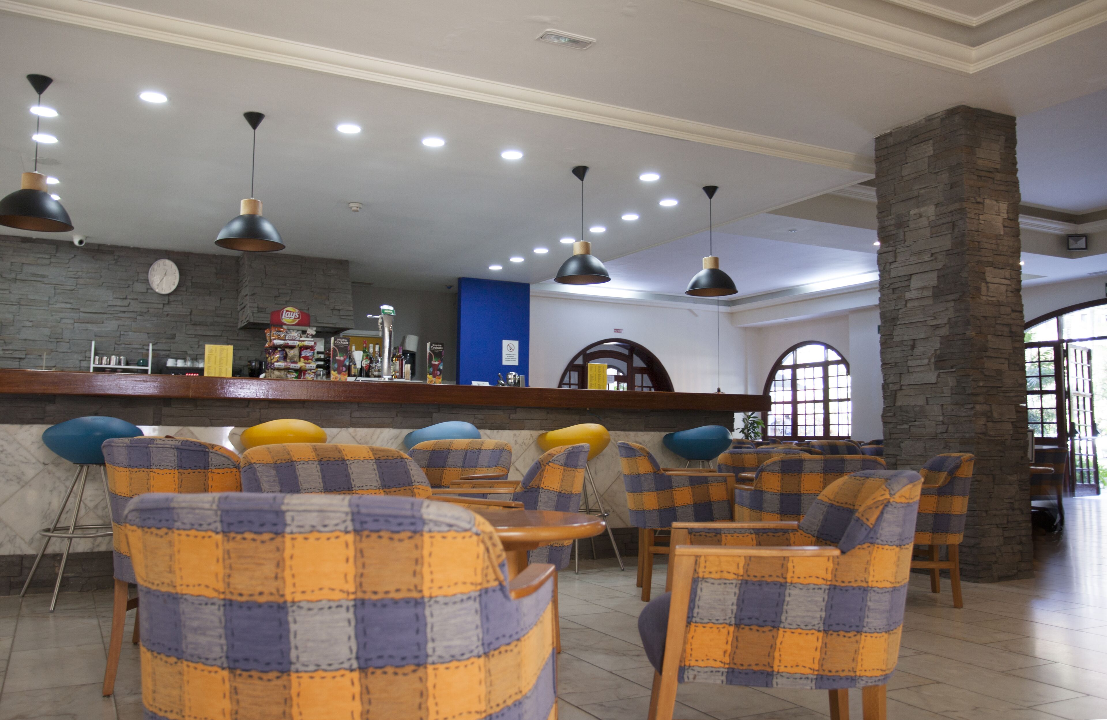 Un restaurante con sillas a cuadros, una barra con un reloj y luminarias colgantes.