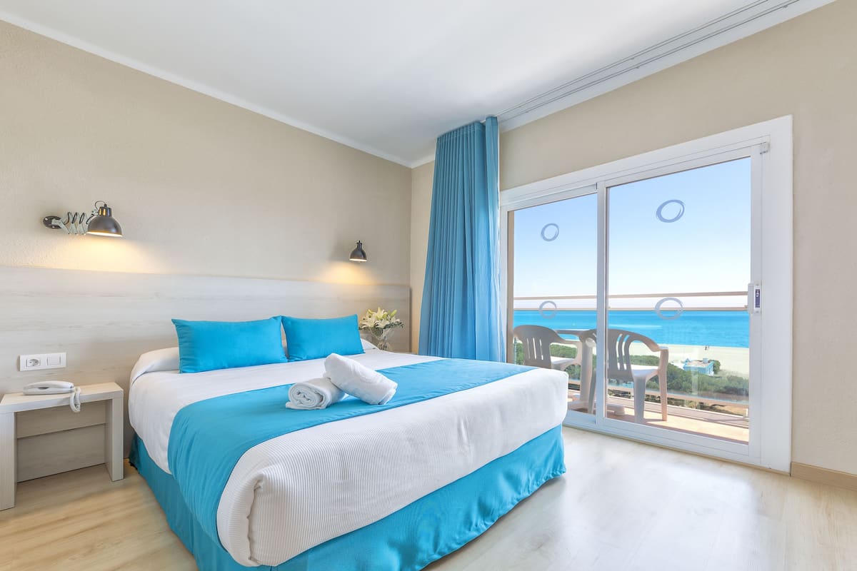 Un dormitorio con cama, mesita de noche, ventana y vista a la playa, además de cortina.
