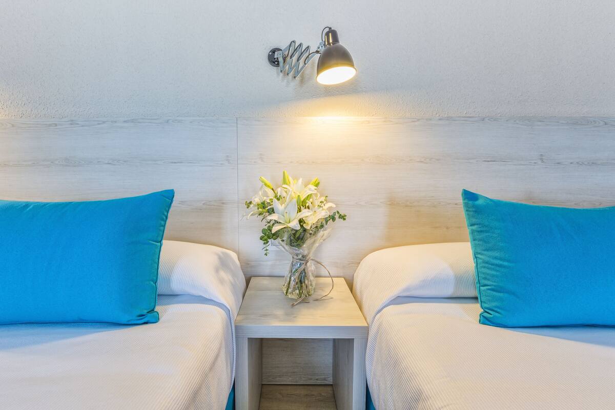 Habitación de hotel con dos camas, cada una con almohada azul y ropa de cama blanca. Un mesita de noche con un florero de flores blancas se encuentra entre las camas.