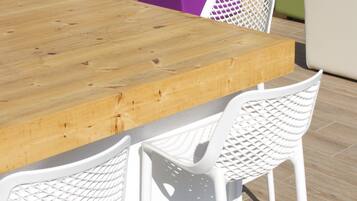 Una mesa de madera con sillas blancas y cojines coloridos en un área de estar al aire libre, luminosa y soleada.