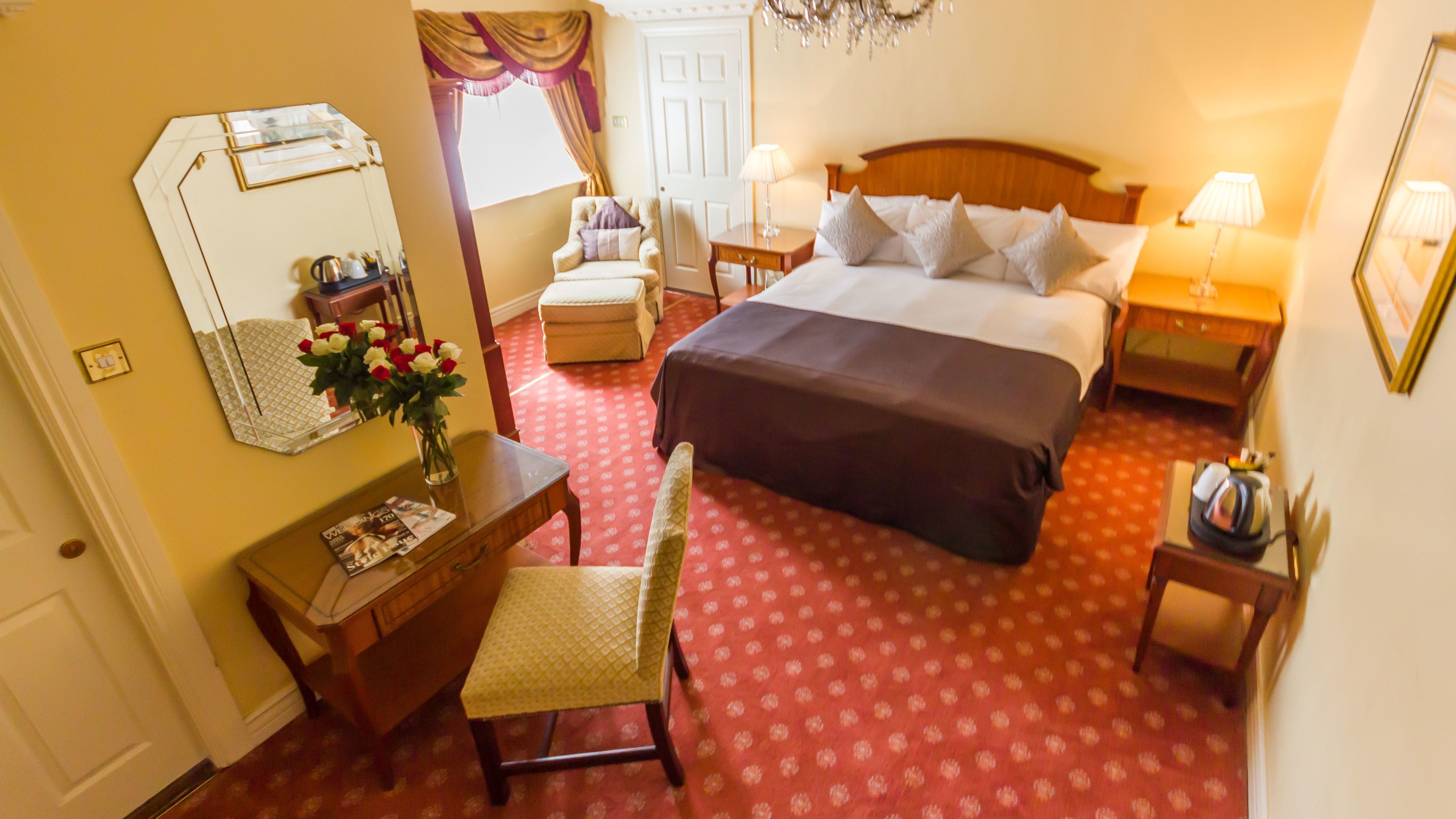 Una camera d'albergo con un letto, una scrivania con una sedia, un piccolo tavolino con una lampada, uno specchio e un vaso con fiori.