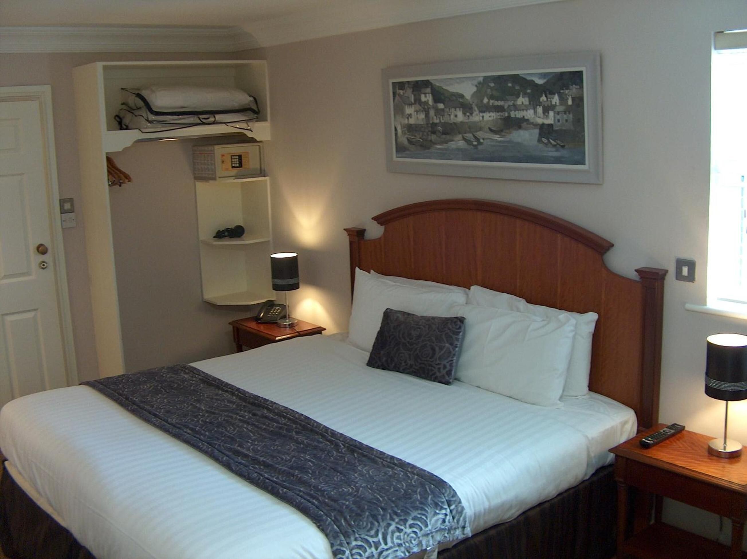 A neatly made bed with a wooden headboard, a bedside table with a lamp, a shelf with folded clothes, and a framed picture on the wall.