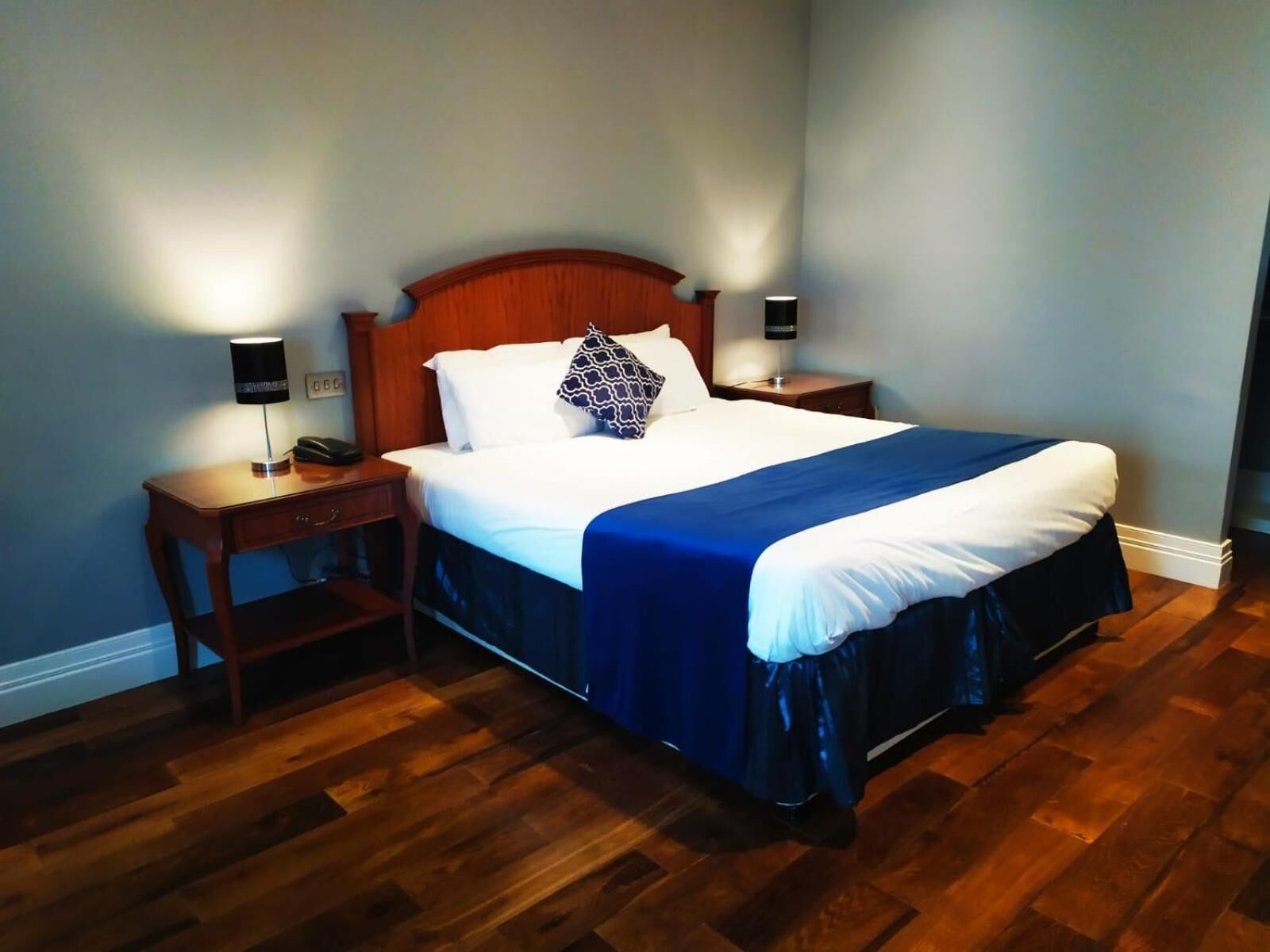 A neatly made bed with a blue bedspread and white linens, flanked by two bedside tables with lamps, in a room with wooden flooring.