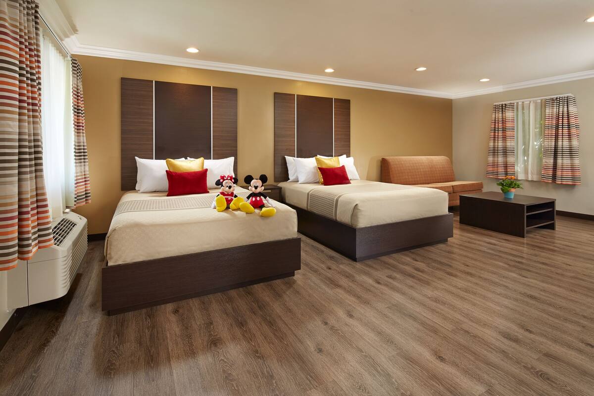 Une chambre d’hôtel avec deux lits, un canapé, une petite table et des peluches à l’effigie de Mickey Mouse sur les lits.