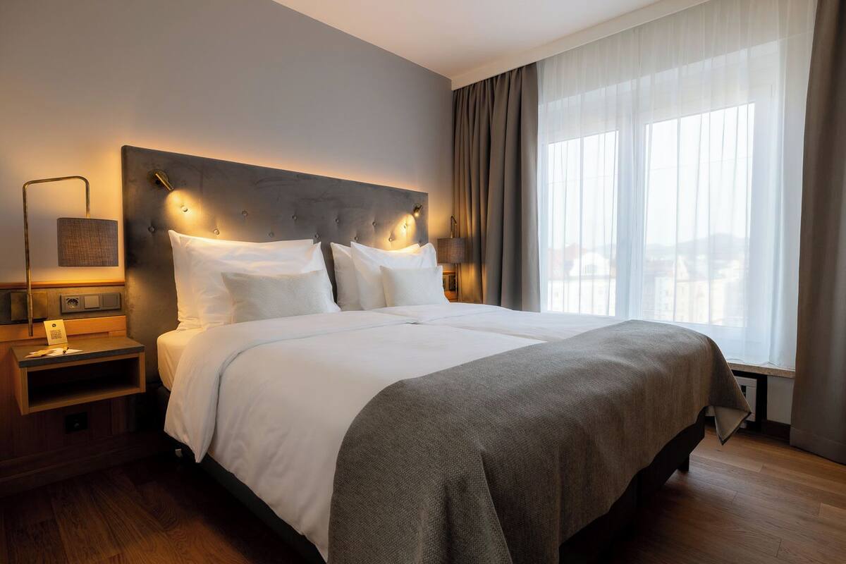 Habitació d'hotel amb un llit gran, una tauleta de nit amb una làmpada, una finestra amb cortines i vistes a edificis a l'exterior.
