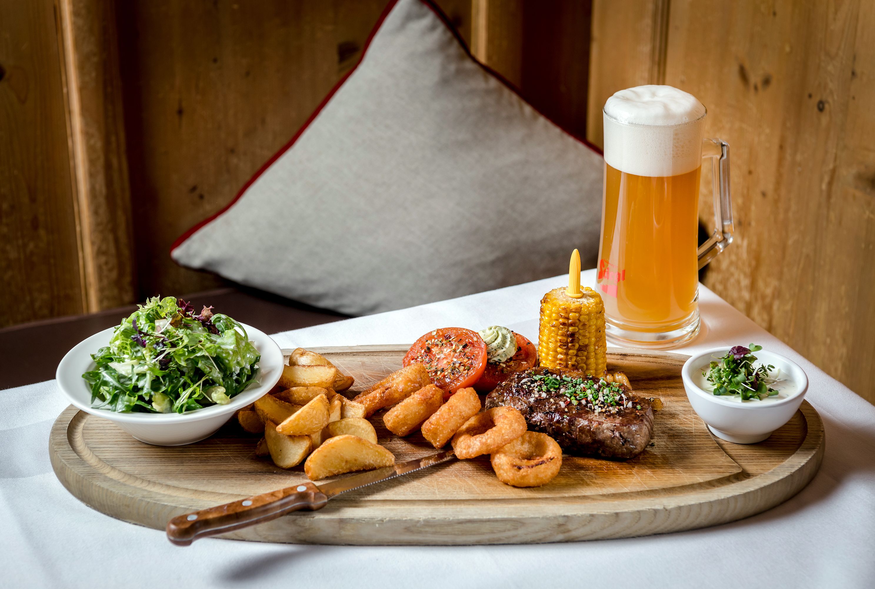 Une table en bois sur laquelle est dressé un repas composé d’une salade, de viande, d’épis de maïs et de chips. On peut également apercevoir une chope de bière.
