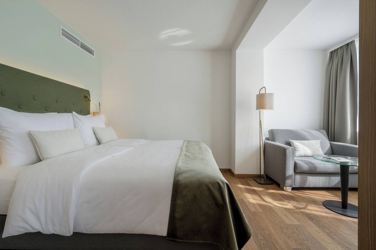 Habitació d'hotel amb un llit, un butaca gris, una taula petita i una làmpada.