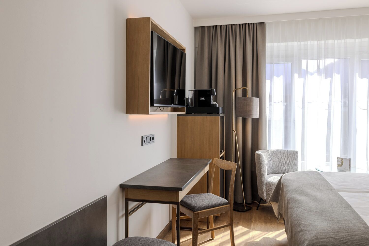 A hotel room with a bed, a desk with a chair, a TV, a lamp, and a window with curtains.