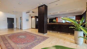 A hotel lobby with a reception desk, a large patterned rug, a potted plant, and a dark pillar.