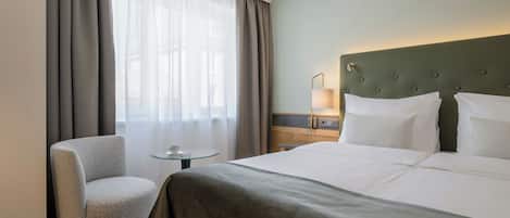 A hotel room with a large bed, a chair, a small table, and a nightstand with a lamp.