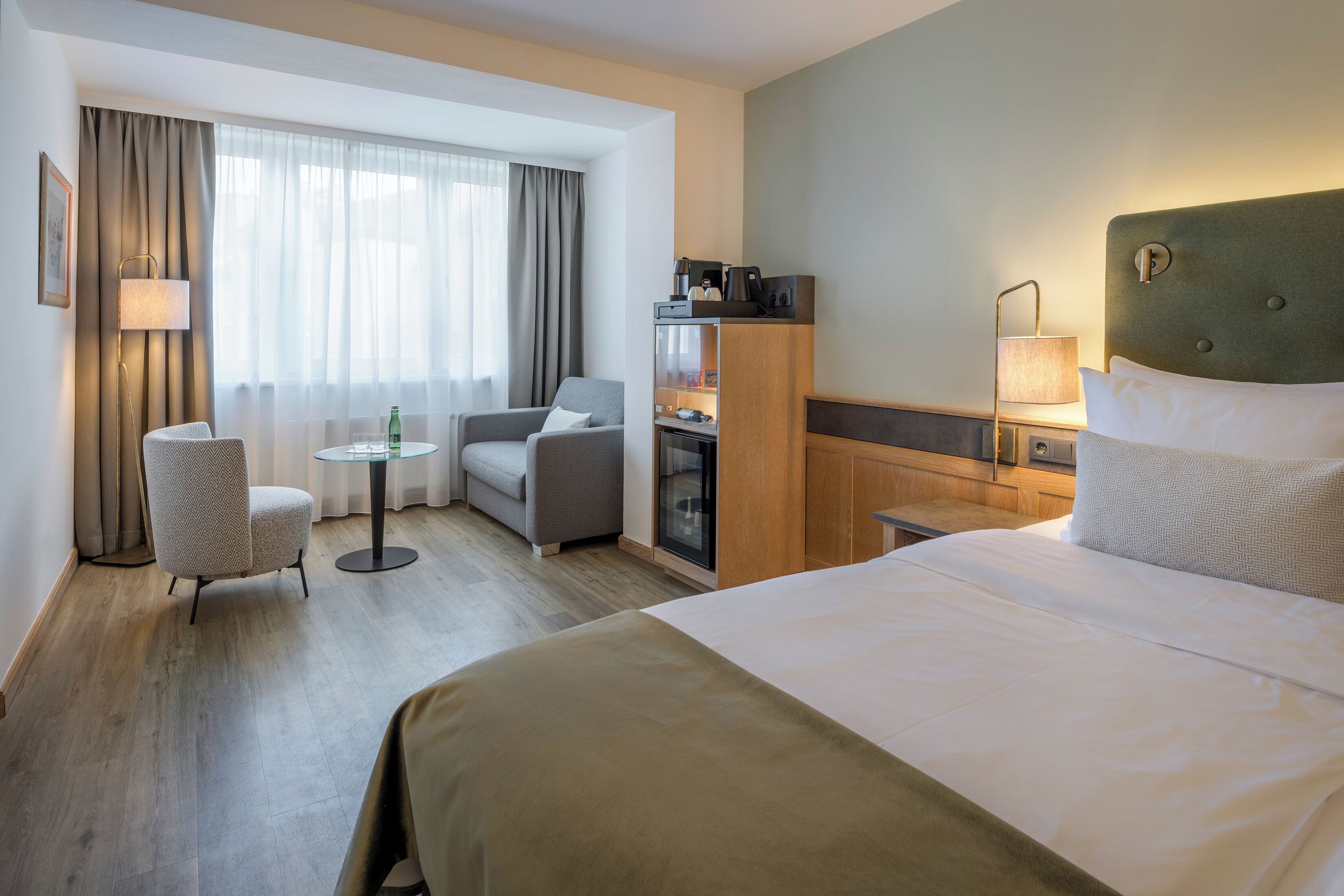 Habitación de hotel con una cama grande, un escritorio, una silla, una mesita, un minibar y una ventana con cortinas.