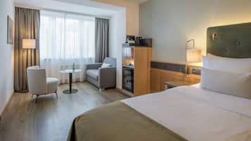 Habitación de hotel con una cama grande, un escritorio, una silla, una mesita, un minibar y una ventana con cortinas.