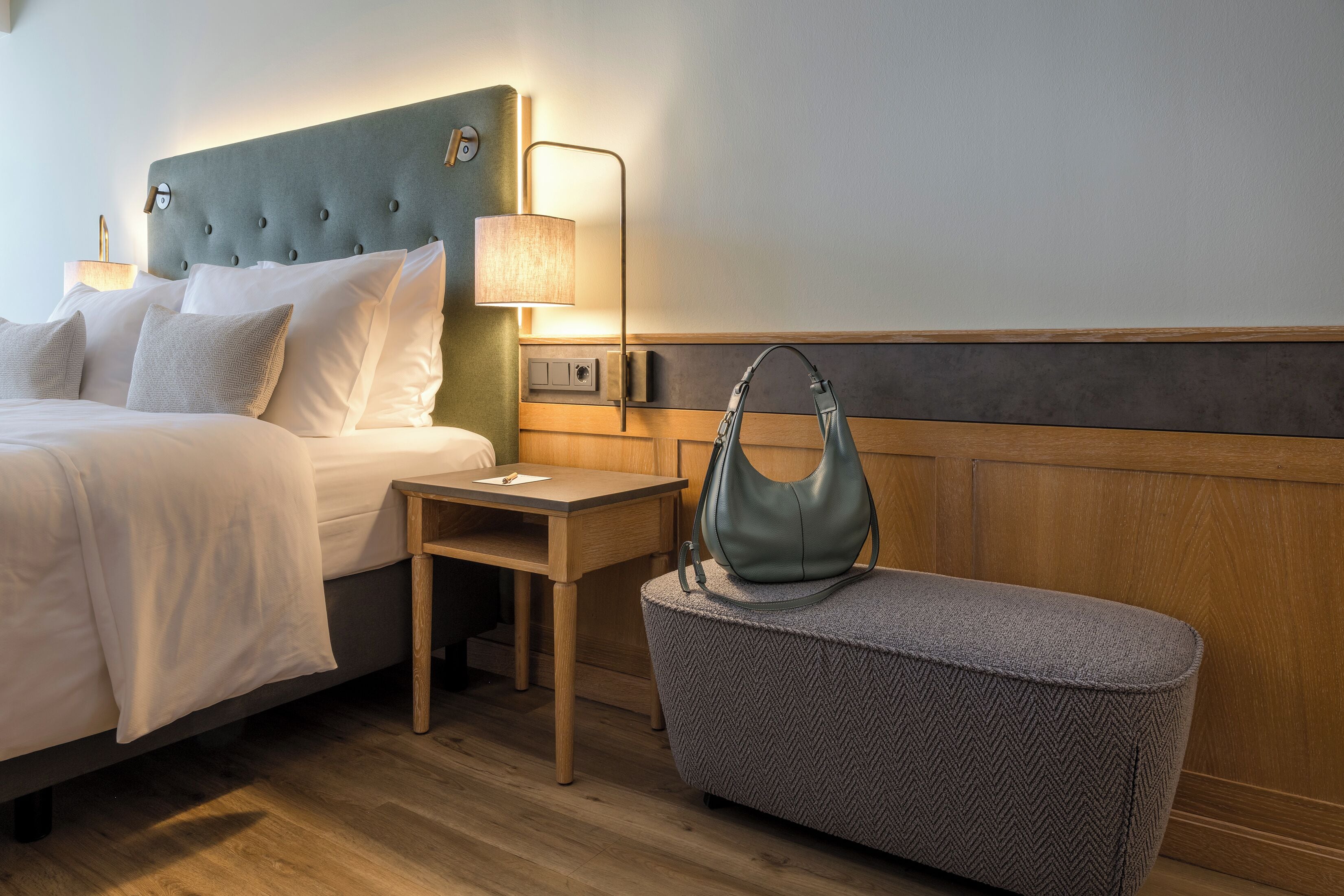 Ein Hotelzimmer mit einem Bett, einem Nachttisch, einer Lampe und einer Handtasche auf einer Bank.