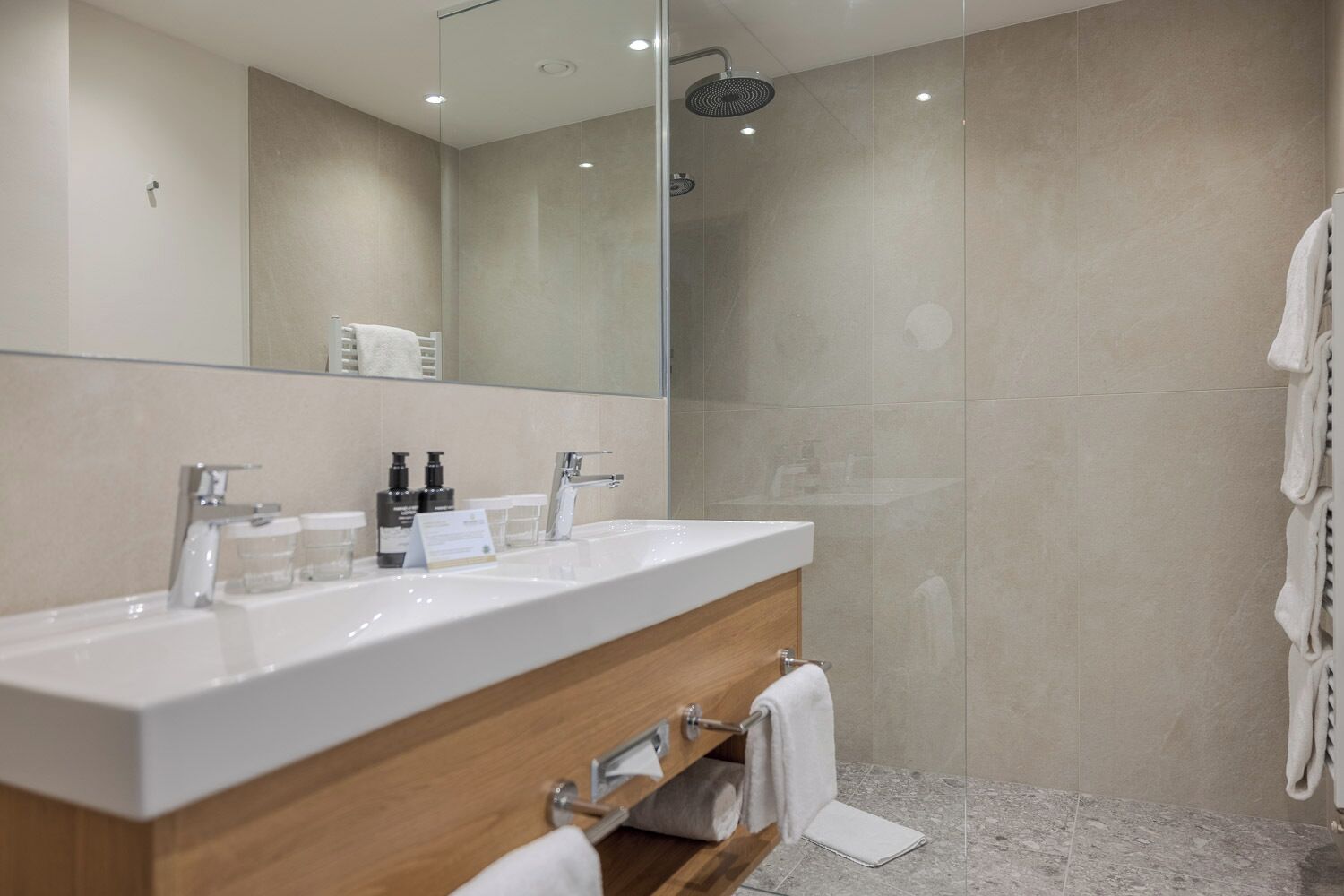 A modern bathroom with a large mirror, two sinks, a shower, and a wooden vanity.