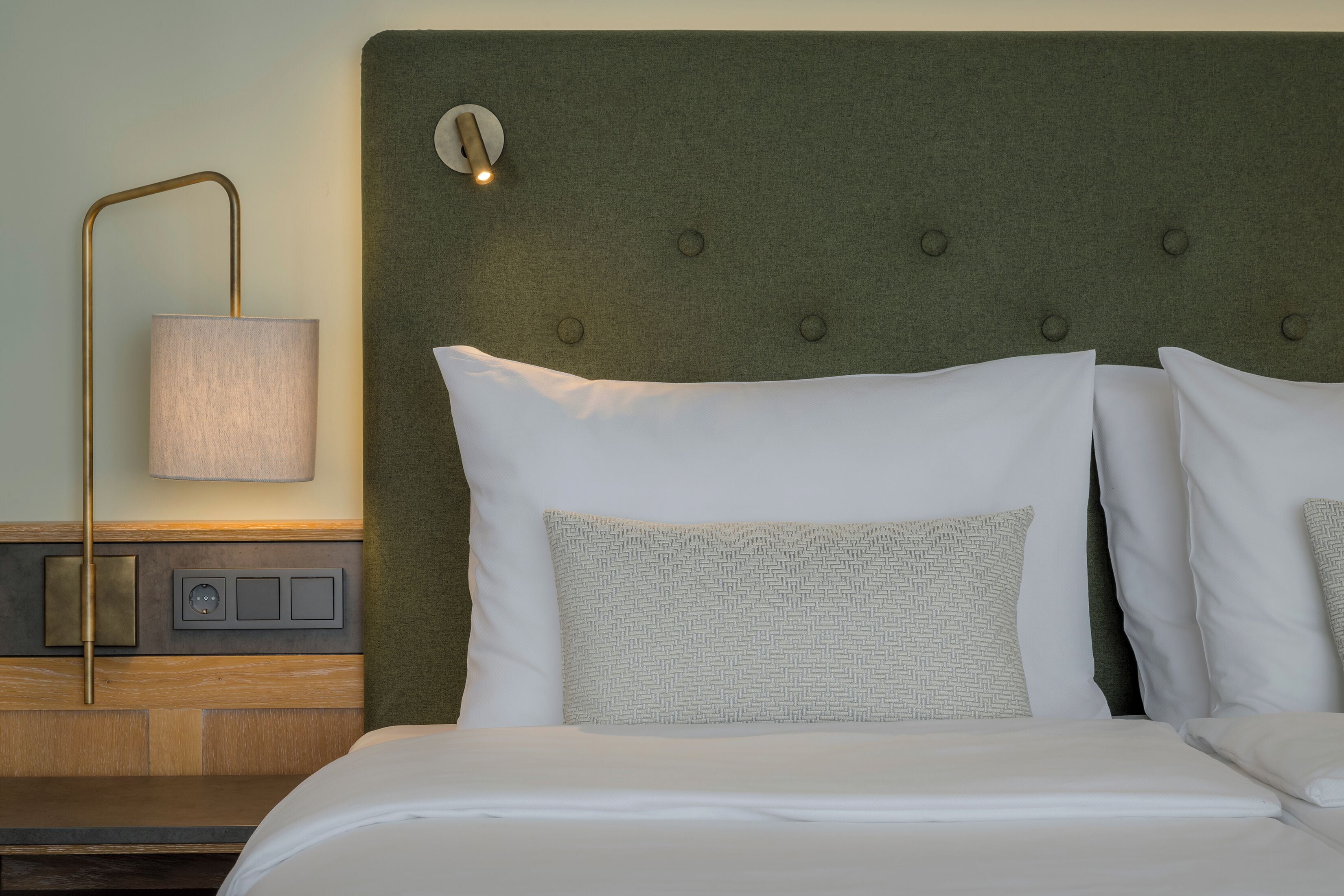 A neatly made bed with white linens and a patterned pillow, a bedside lamp, and a wall-mounted light fixture.