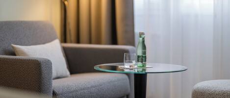 A modern living room with a grey sofa, a glass table with a bottle and glass, and a lamp in the background.
