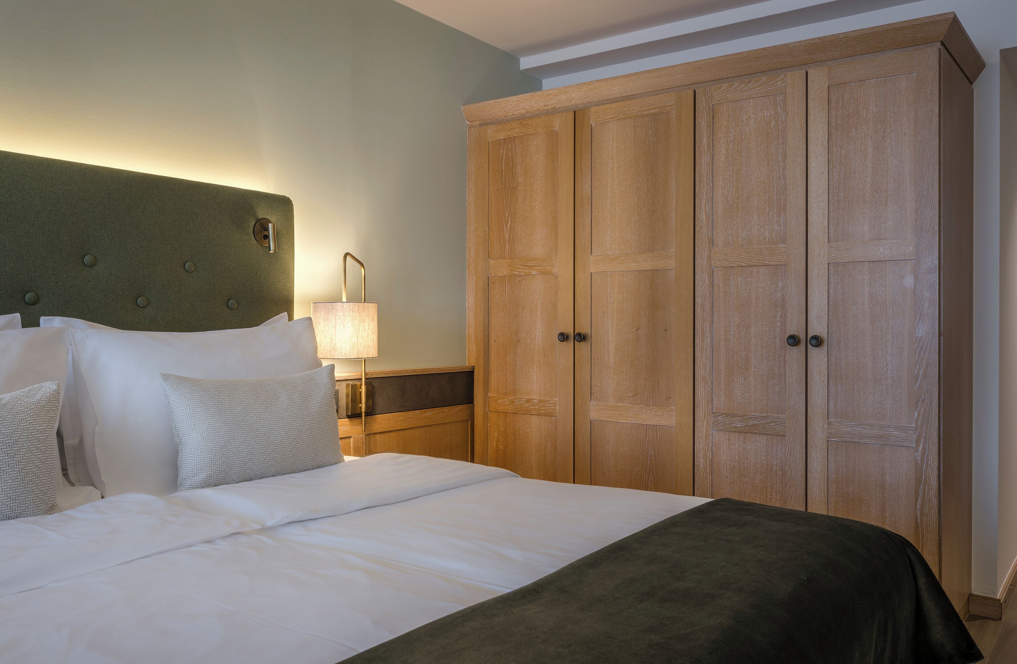 Una cama bien hecha con sábanas blancas y un plaid oscuro, un armario de madera con dos puertas y una mesita de noche con una lámpara.