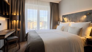 Une chambre d’hôtel équipée d’un lit, d’un bureau, d’une chaise et offrant une vue sur les bâtiments à travers de grandes fenêtres.