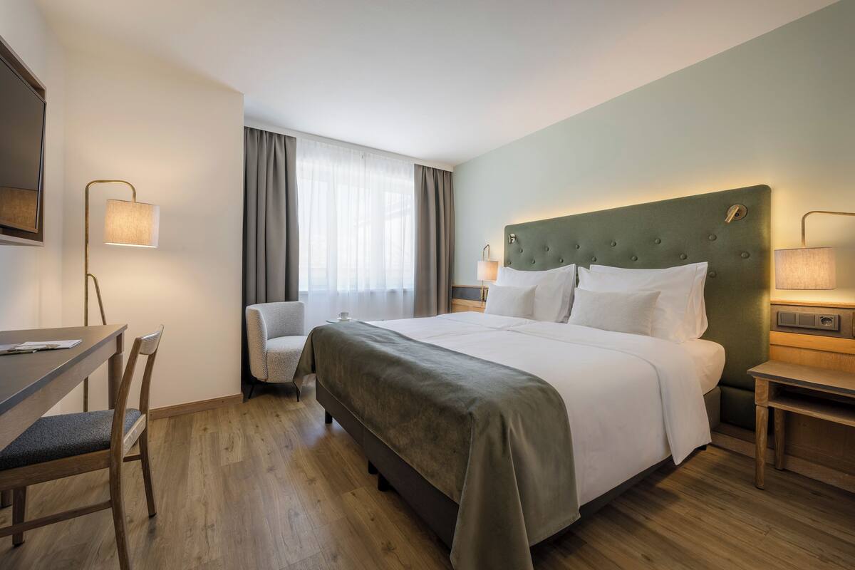 Habitació d'hotel amb un llit gran, un escriptori amb una cadira, una cadira addicional, una làmpada i una finestra amb cortines.