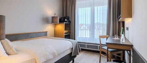 A hotel room with a bed, a desk, a chair, a lamp, a heater, and a window with curtains.