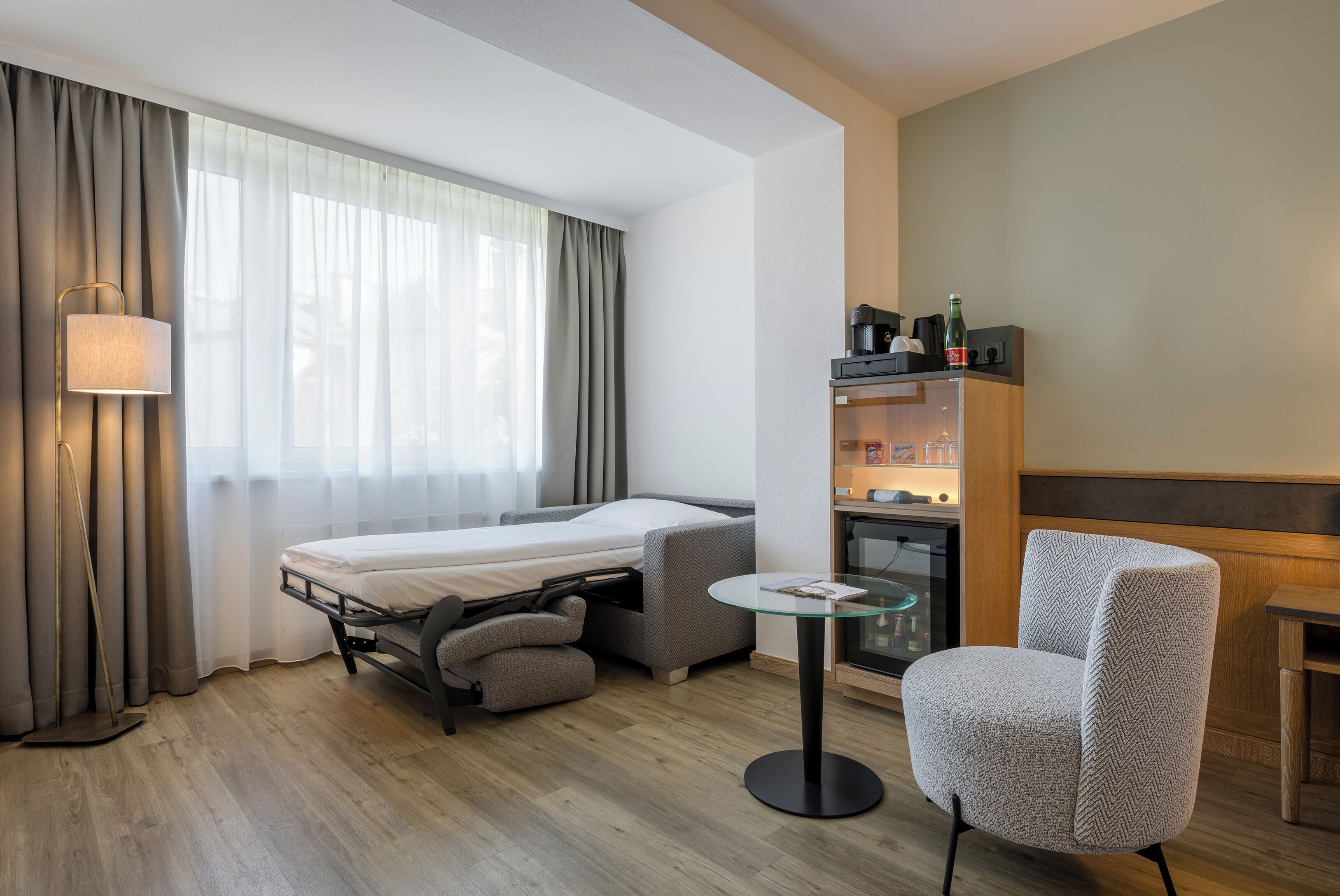 A compact hotel room with a bed, a small sofa, a glass table, a chair, a bookshelf, and a fireplace.