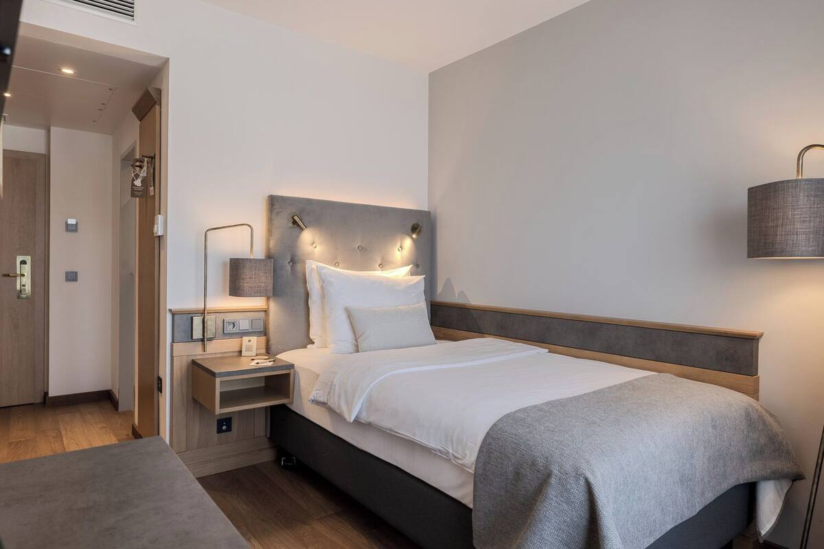 Una habitació d'hotel moderna amb un llit, una taula de nit, una làmpada i una porta que condueix a una altra habitació.