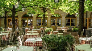 Un café en plein air avec des tables et des chaises, entouré d’arbres et bordé d’un bâtiment aux fenêtres en arc de cercle.