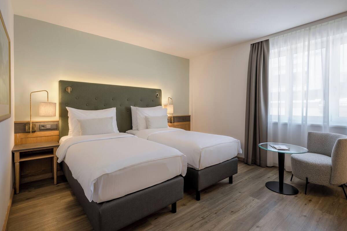 Habitació d'hotel amb dos llits, una taula petita, una cadira i una petita taula de nit amb làmpades.