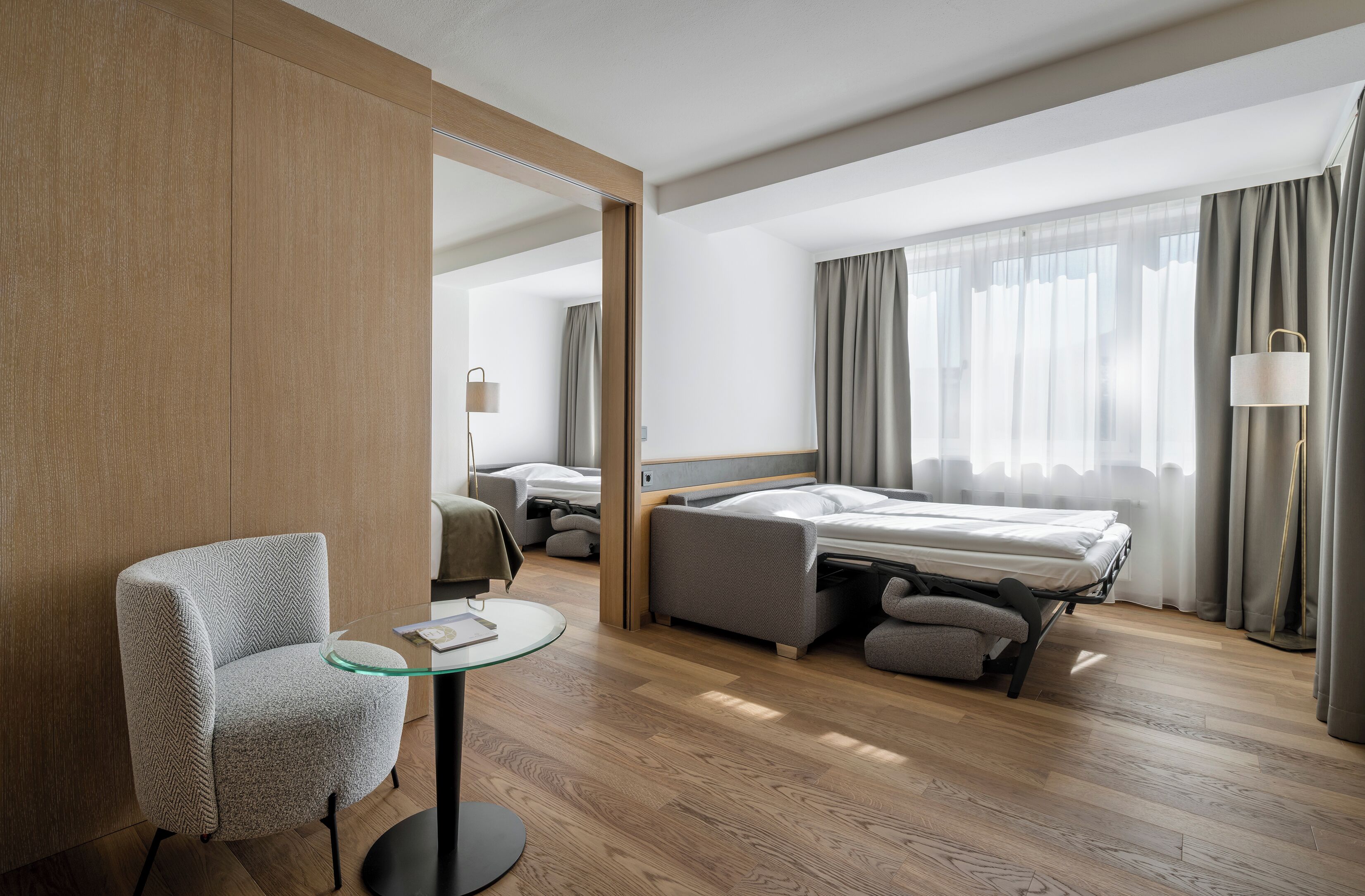 A modern hotel room with a bed, a seating area, a glass table, and a large mirror.