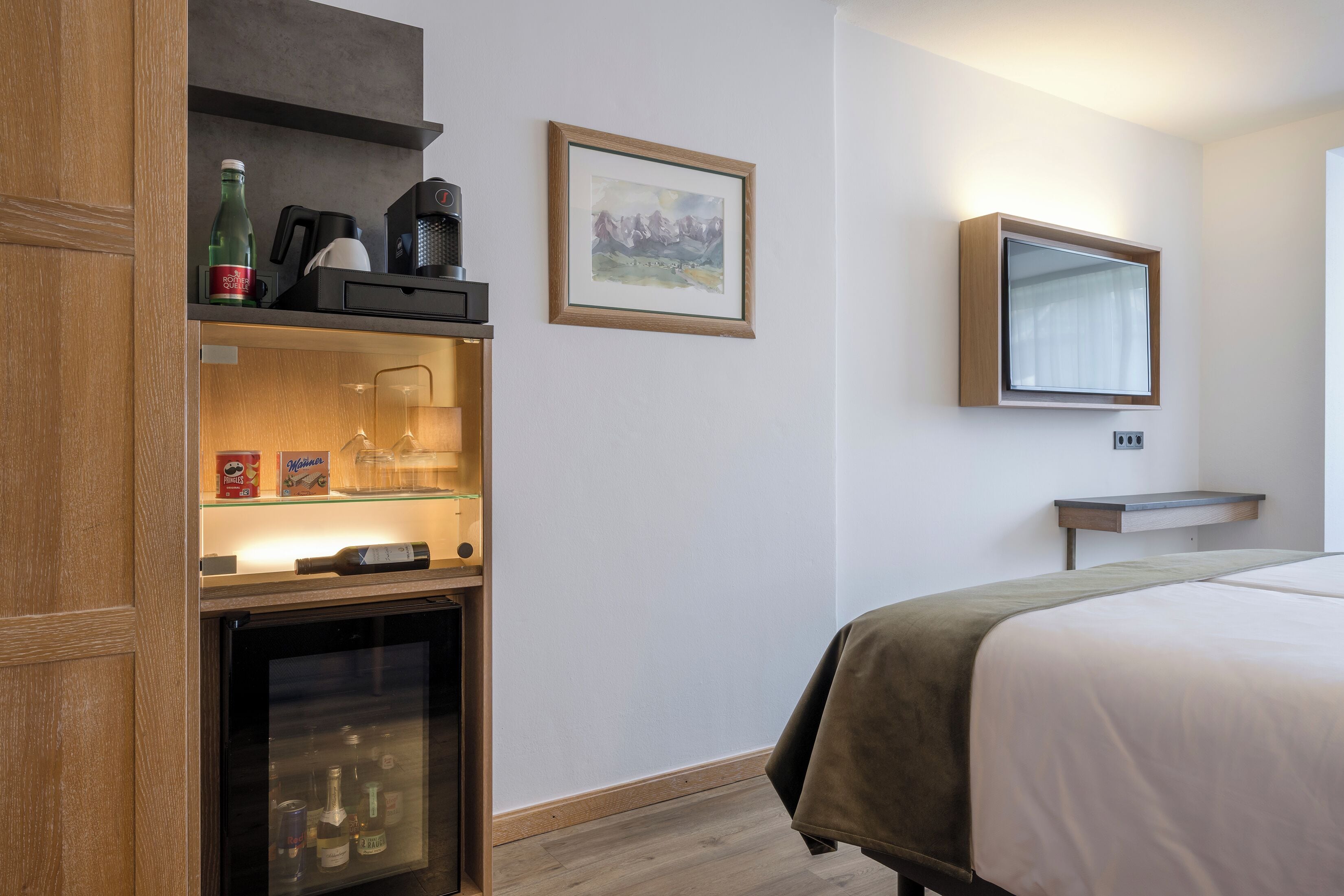 A hotel room with a bed, a minibar, a coffee machine, a framed picture, and a mirror.