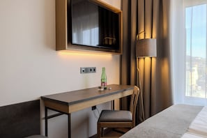 Una habitación de hotel con una cama, un escritorio con silla, un televisor montado en la pared, una lámpara y una ventana con vistas.