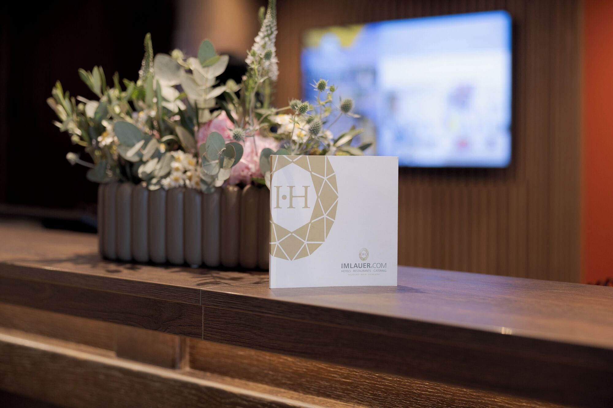 Un comptoir d’accueil agrémenté d’un arrangement floral et d’une enseigne affichant le logo et le nom de l’hôtel.