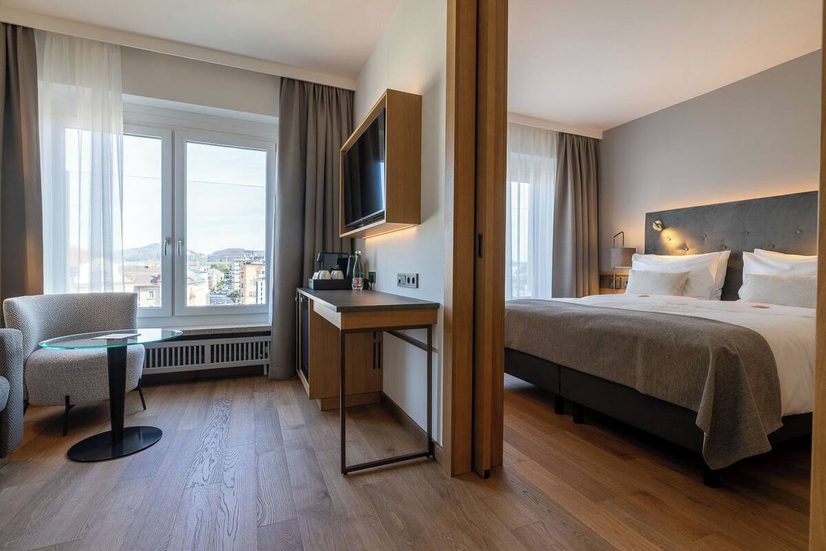 Habitació d'hotel amb llit, escriptori, cadira, televisió i finestres grans.