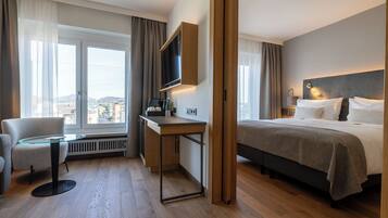 Habitación de hotel con cama, escritorio, silla, televisor y ventanas amplias.
