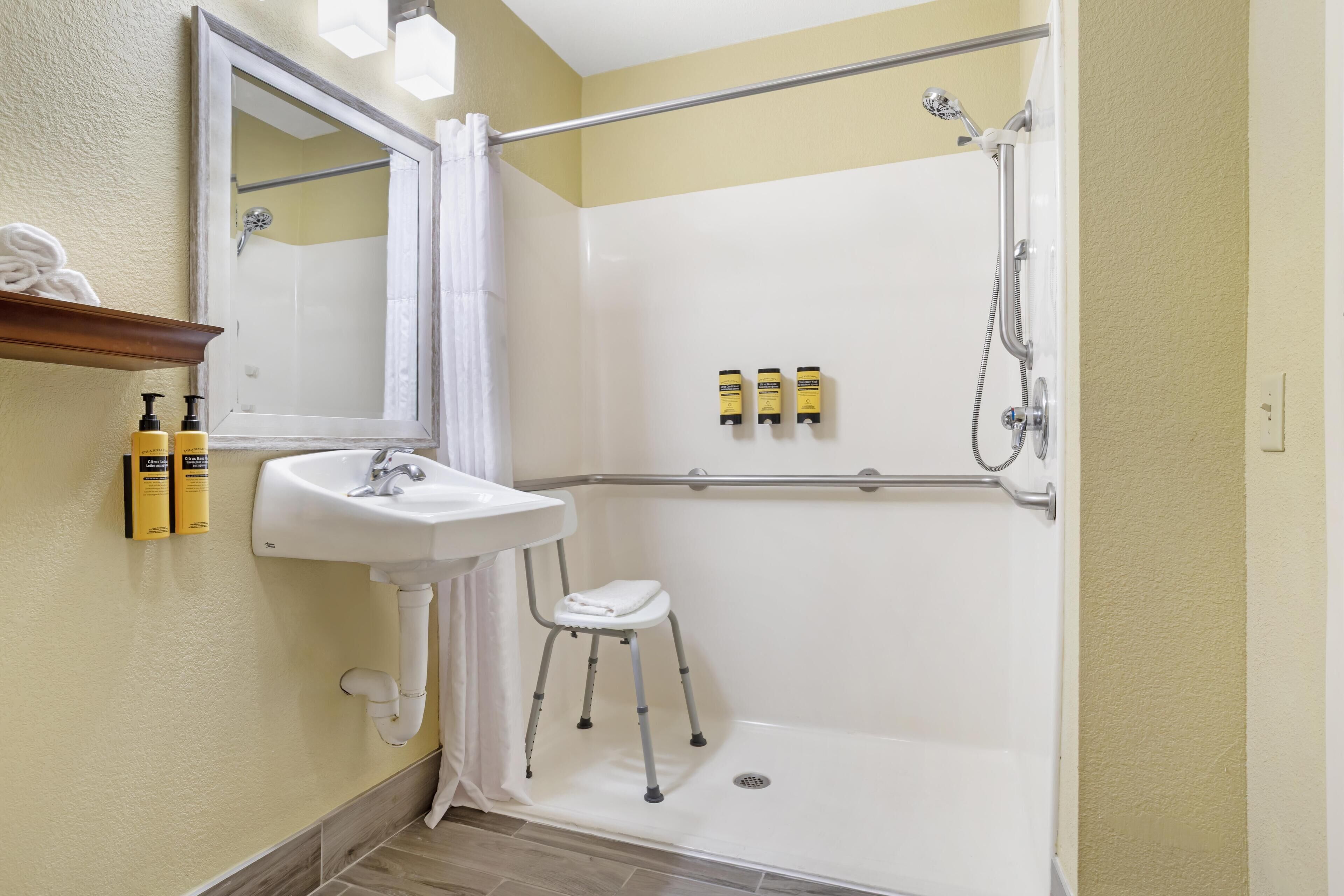 Un baño con lavamanos blanco, espejo, ducha con barra de apoyo y un taburete pequeño.