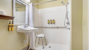 Un baño con lavamanos blanco, espejo, ducha con barra de apoyo y un taburete pequeño.
