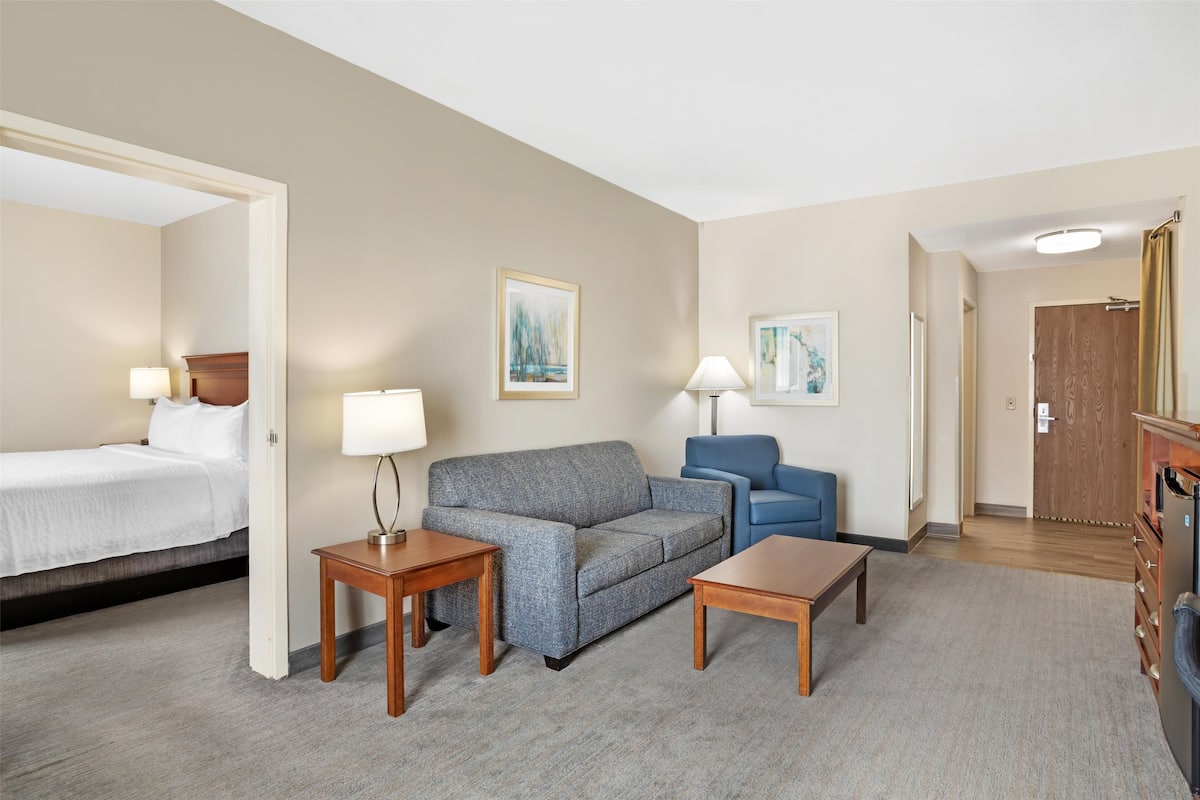 Een hotelkamer met een bed, een grijze bank, een blauwe fauteuil, twee houten bijzettafeltjes en een houten kledingkast.