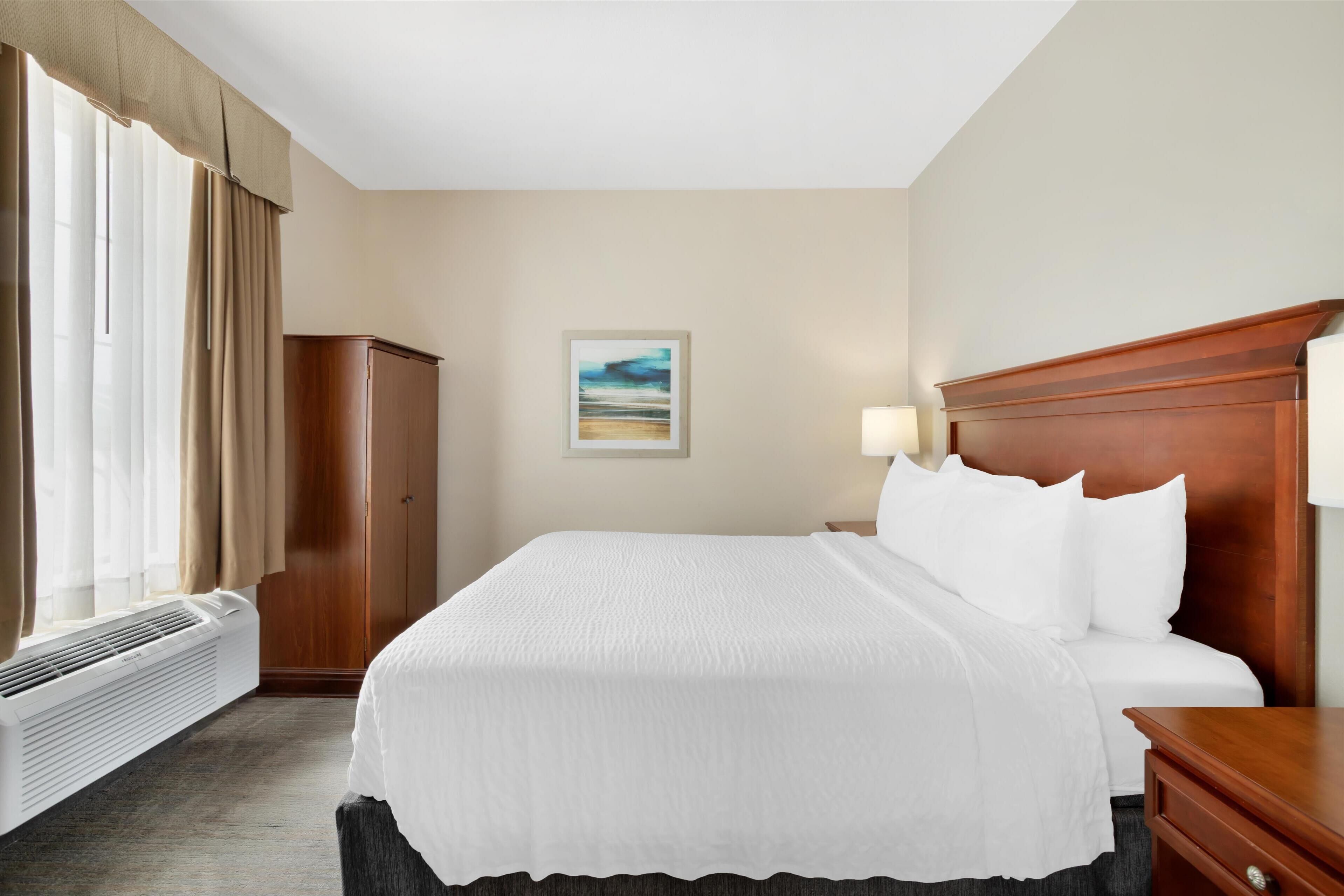 Habitación de hotel con una cama grande, cabecera de madera, mesita de noche, lámpara, armario y ventana con cortinas.