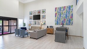 Un vaste espace de vie comprenant un canapé gris, deux fauteuils, une table basse et un téléviseur fixé au mur. Des œuvres d’art encadrées et une peinture abstraite aux couleurs vives ornent le mur.